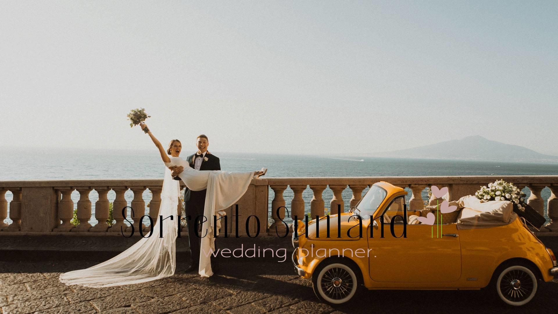 Sorrento Town. Bride is in grooms arms facing the camera. The bride has her bouquet in hand with her armed raised celebrating their recent commitment. Her vail flows to the ground and so does the train of her white wedding dress. They stand next to a yellow vintage convertible Fiat 500 that has a flowers composition laying on it. In the background is the ocean and the lightly foggy sky, which shows an outline of a mountain. Text goes across the bottom of the image reading “Sorrento Sunland wedding planner.”